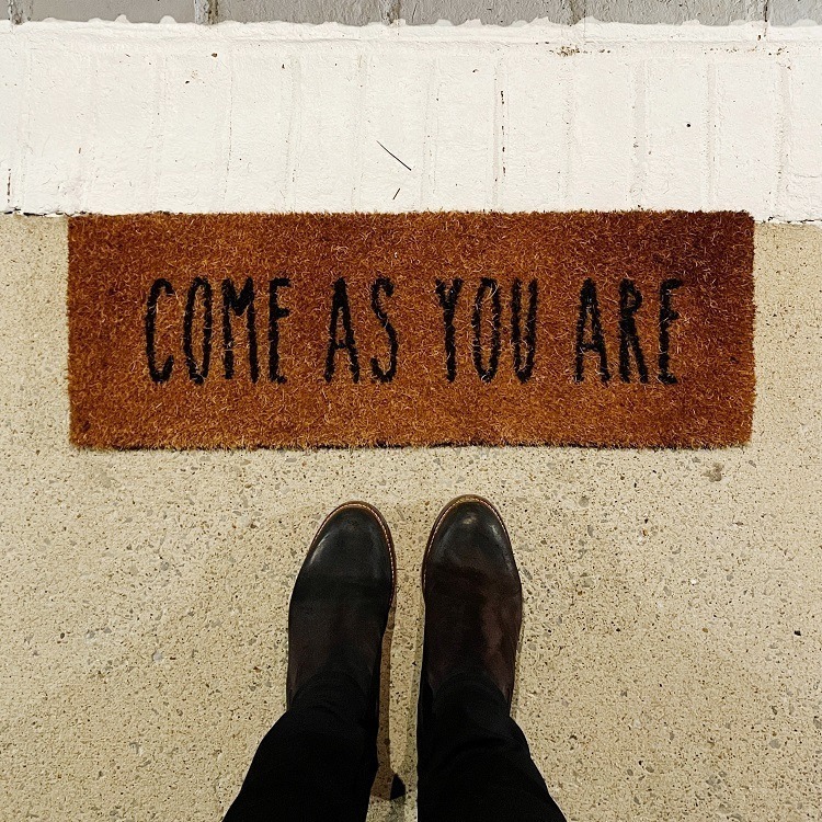Two feet in black shoes are standing next to a door mat with the phrase 'Come as you are' on it.