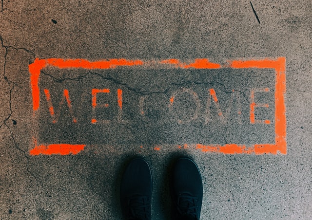 Two feet in black shoes are standing next to the word 'welcome', painted in orange on a concrete floor.