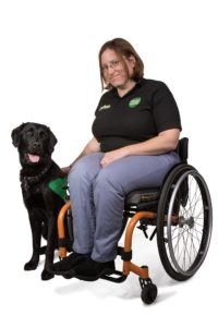 Liz in a wheelchair with a black Labrador sat to the left of her, its tongue out.
