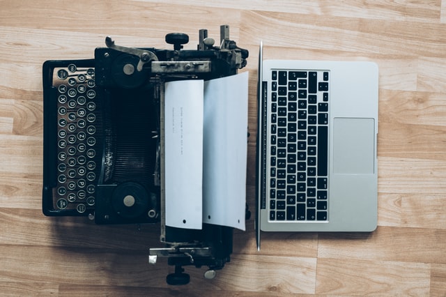 A black typewriter with a white sheet of paper in it, back-to-back with a grey laptop.